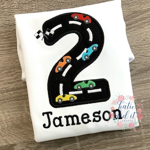 May include: White onesie with a black number two embroidered on it. The number two is shaped like a race track with colorful cars driving around it. The onesie is personalized with the name "Jameson".