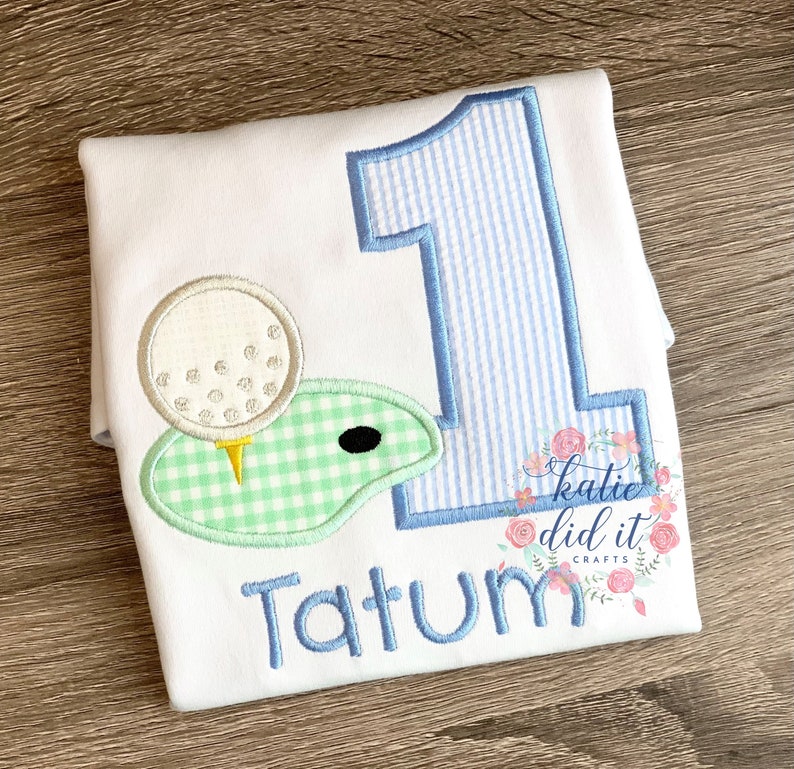 Shirt with applique "1" using light blue seersucker fabric. Next to the "1" is an applique with white golf ball and mint green gingham grass. Child's name is optionally below the design.