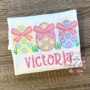 May include: White t-shirt with three embroidered pastel Easter eggs with pink bows and the name "Victoria" embroidered below. The text "katie did it crafts" is in the bottom right corner.