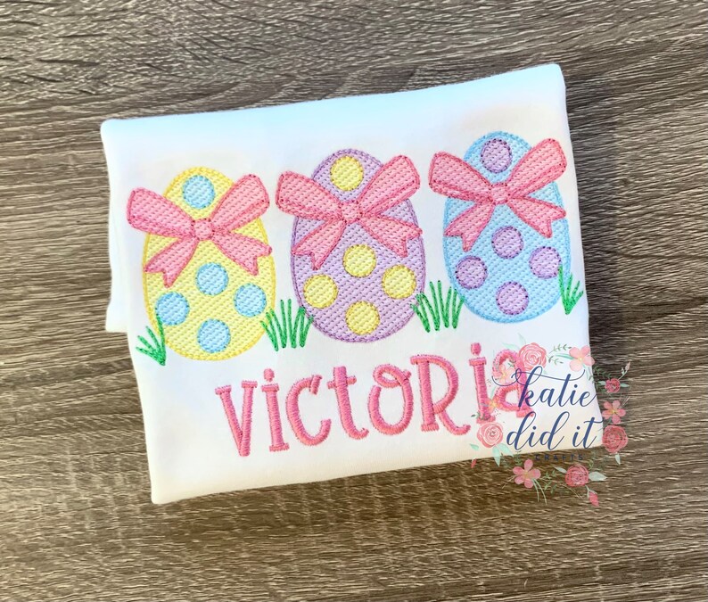 May include: White t-shirt with three embroidered pastel Easter eggs with pink bows and the name "Victoria" embroidered below. The text "katie did it crafts" is in the bottom right corner.
