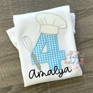 May include: White t-shirt with a blue and white checkered number four, a chef's hat, and a whisk. The name "Amaya" is embroidered below the number.