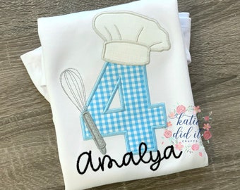 Op de afbeelding: Wit T-shirt met een blauw-wit geruit nummer vier, een koksmuts en een garde. De naam "Amaya" is onder het nummer geborduurd.