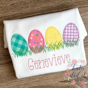 Puede incluir: Una camiseta blanca con cuatro huevos de Pascua bordados en diferentes patrones y colores. Los huevos est&aacute;n alineados en una fila y est&aacute;n sentados sobre una cama de hierba verde bordada. El nombre "Genevieve" est&aacute; bordado debajo de los huevos en hilo rosa.