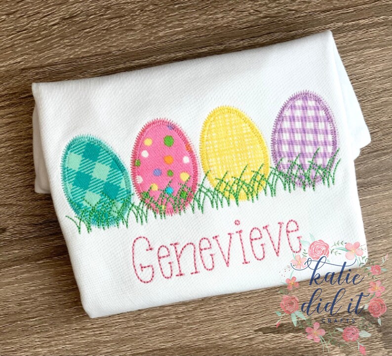 Puede incluir: Una camiseta blanca con cuatro huevos de Pascua bordados en diferentes patrones y colores. Los huevos est&aacute;n alineados en una fila y est&aacute;n sentados sobre una cama de hierba verde bordada. El nombre "Genevieve" est&aacute; bordado debajo de los huevos en hilo rosa.