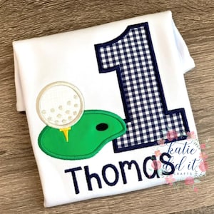 Pu&ograve; includere: Tutina bianca con un numero uno in vichy blu e bianco, un campo da golf verde con una palla da golf bianca e il nome "Thomas" in ricamo blu.