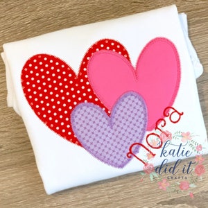 May include: Three felt hearts in red, pink, and purple with white polka dots, crosshatch, and solid patterns. The hearts are layered on top of each other. The text "Nora" is stitched in red thread below the hearts. The text "katie did it crafts" is in the bottom right corner.