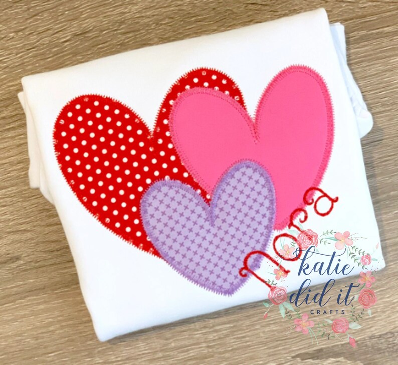May include: Three felt hearts in red, pink, and purple with white polka dots, crosshatch, and solid patterns. The hearts are layered on top of each other. The text "Nora" is stitched in red thread below the hearts. The text "katie did it crafts" is in the bottom right corner.