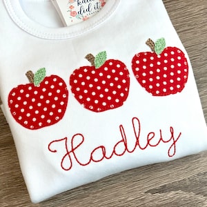 May include: White t-shirt with three red apples with white polka dots and a green leaf on each. The name "Hadley" is embroidered in red below the apples.