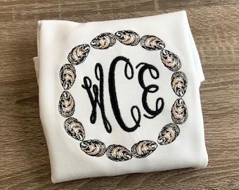 以下が含まれることがあります： 白いTシャツに、黒い刺繍で「WCE」のモノグラムが施され、その周りをオイスターの貝殻の円が囲んでいます。