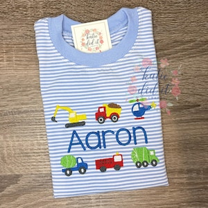 May include: Blue and white striped t-shirt with a light blue collar. The shirt features embroidered images of construction vehicles and a helicopter. The name "Aaron" is embroidered in blue letters. A tag that says "katie did it" is attached.