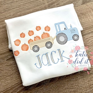 May include: A white onesie with a blue tractor pulling a wagon full of orange pumpkins. The text "JACK" is embroidered in blue and yellow thread below the tractor. The text "katie did it crafts" is embroidered in black thread in the lower right corner.