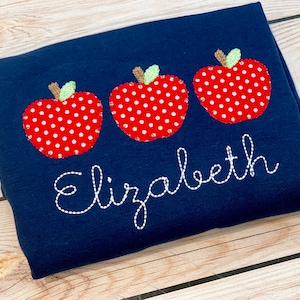 May include: A navy blue t-shirt with three red and white polka dot apples and the name "Elizabeth" embroidered in white thread.