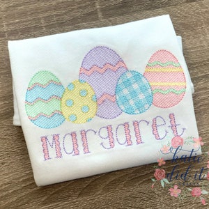 K&ouml;nnte beinhalten: Ein wei&szlig;es besticktes Shirt mit einer Reihe von f&uuml;nf pastellfarbenen Ostereiern und dem Namen "Margaret" darunter gestickt.