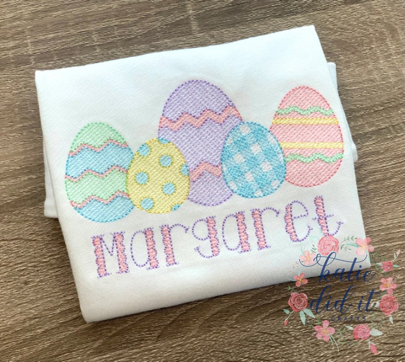 K&ouml;nnte beinhalten: Ein wei&szlig;es besticktes Shirt mit einer Reihe von f&uuml;nf pastellfarbenen Ostereiern und dem Namen "Margaret" darunter gestickt.
