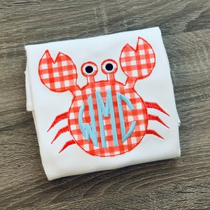 May include: White babygrow with a red and white gingham crab applique. The crab has a blue monogram in the centre.