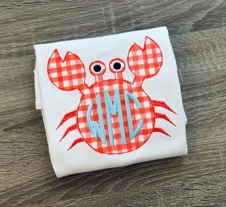 May include: White babygrow with a red and white gingham crab applique. The crab has a blue monogram in the centre.