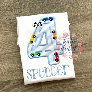 以下が含まれることがあります： 白いTシャツに青い数字の4が刺繍されています。数字の4はレースコースの形をしていて、小さな車がその周りを走っています。数字の下には「Spencer」という名前が刺繍されています。