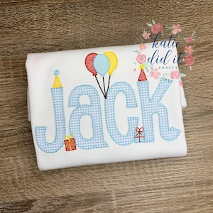May include: White t-shirt with the name "Jack" embroidered in blue gingham letters. The design includes party hats, balloons, and a gift box, perfect for a birthday celebration. The text "katie did it crafts" is also visible.