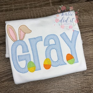 May include: White t-shirt with the name "GRAY" in blue gingham letters, bunny ears, and Easter eggs. The shirt has a playful design with colorful accents, perfect for Easter or spring. The shirt is folded on a wooden surface.