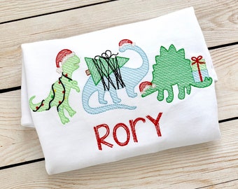 May include: A white t-shirt with an embroidered design of three dinosaurs wearing Santa hats. The dinosaurs are carrying a Christmas tree and a gift. The name "Rory" is embroidered below the dinosaurs.