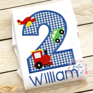 May include: A white onesie with a blue and white gingham number two. The number two is decorated with a red airplane, a green car, and a red train. The name "William" is embroidered below the number.