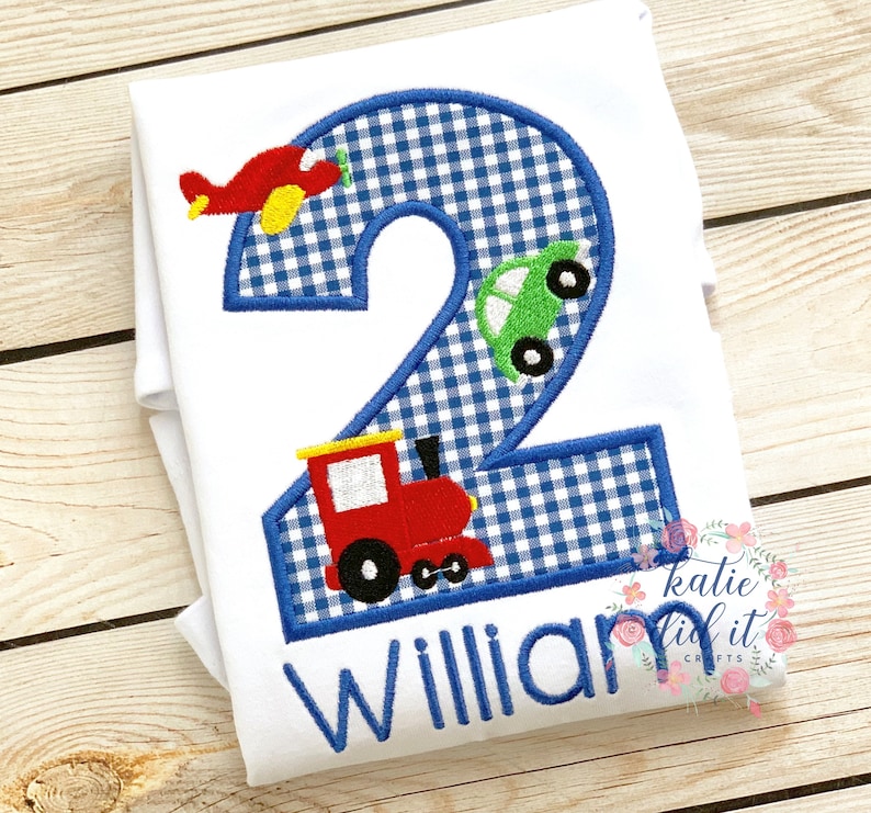 May include: A white onesie with a blue and white gingham number two. The number two is decorated with a red airplane, a green car, and a red train. The name "William" is embroidered below the number.