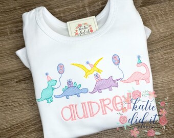 Puede incluir: Camiseta blanca de manga corta con bordado de dinosaurios rosas. Los dinosaurios llevan gorros de fiesta y sostienen globos y regalos. La camiseta también tiene el nombre "Audre" y el texto "katie did it crafts" bordado.