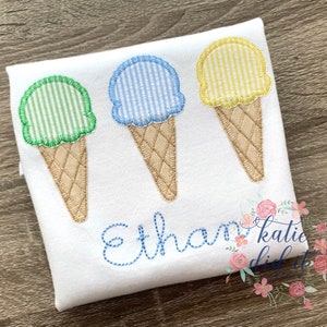 Op de afbeelding: Drie geborduurde ijshoorns op een wit shirt. De hoorns zijn groen, blauw en geel met witte strepen. Het shirt heeft de naam "Ethan" geborduurd in blauwe draad.