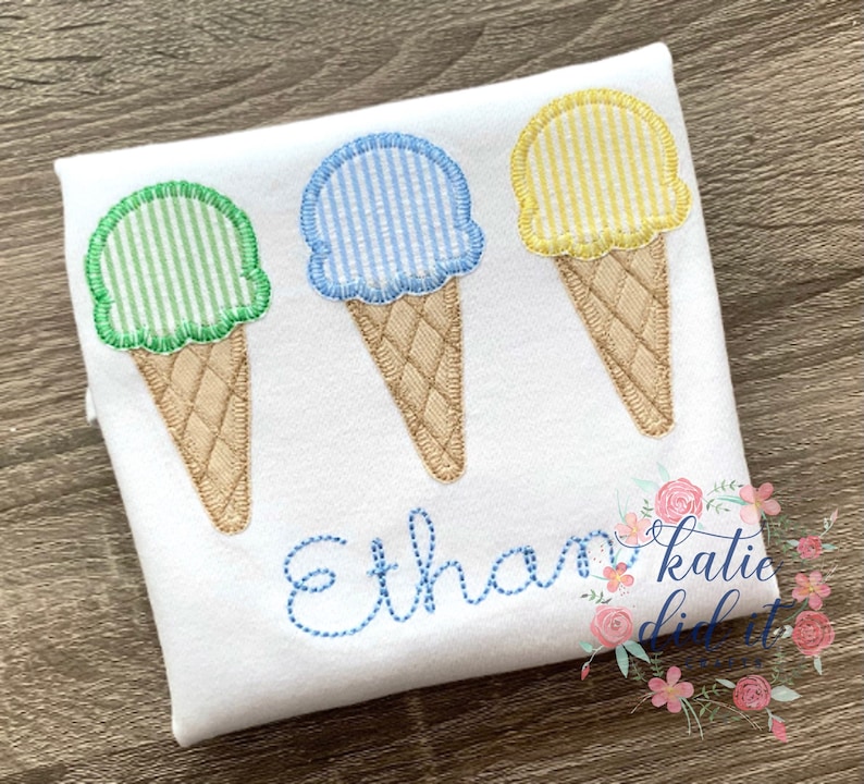 Op de afbeelding: Drie geborduurde ijshoorns op een wit shirt. De hoorns zijn groen, blauw en geel met witte strepen. Het shirt heeft de naam "Ethan" geborduurd in blauwe draad.