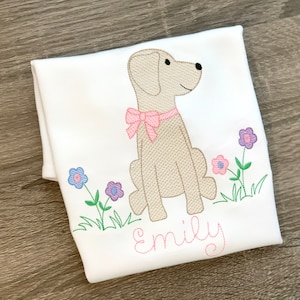 May include: A white t-shirt with an embroidered image of a beige dog wearing a pink bow and sitting in a field of green grass with purple and pink flowers. The name "Emily" is embroidered in pink below the dog.