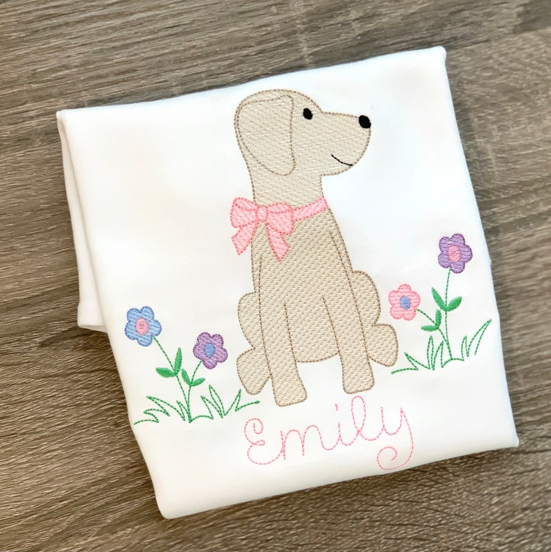 May include: A white t-shirt with an embroidered image of a beige dog wearing a pink bow and sitting in a field of green grass with purple and pink flowers. The name "Emily" is embroidered in pink below the dog.