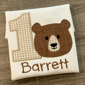 Op de afbeelding: Witte romper met een bruin berenhoofd, een geruit nummer &eacute;&eacute;n en de naam "Barrett" geborduurd in bruin.