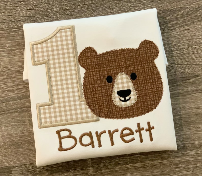 Op de afbeelding: Witte romper met een bruin berenhoofd, een geruit nummer &eacute;&eacute;n en de naam "Barrett" geborduurd in bruin.