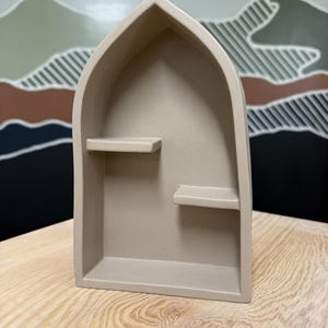 May include: A beige, arch-shaped wall shelf with a pointed top and two small, staggered internal shelves. The structure is made of a solid, matte-finished material and features a deep, recessed interior space for displaying small items.
