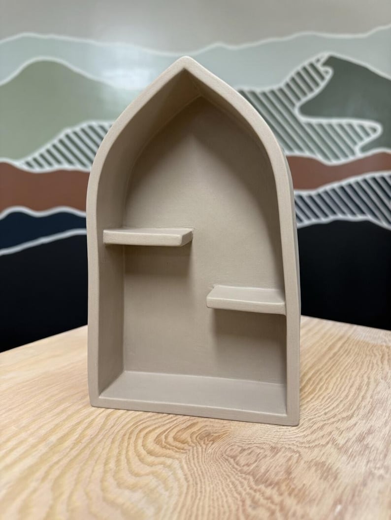 May include: A beige, arch-shaped wall shelf with a pointed top and two small, staggered internal shelves. The structure is made of a solid, matte-finished material and features a deep, recessed interior space for displaying small items.