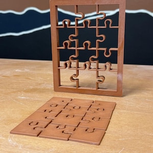 May include: A brown, rectangular puzzle frame standing upright with interlocking puzzle pieces inside, and a matching set of twelve flat, square puzzle pieces arranged in a grid on the surface in front of the frame.