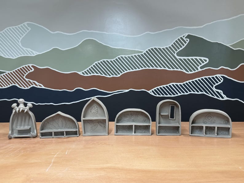 May include: A set of six gray ceramic miniature shelves with various architectural shapes, including arched, pointed, and rounded designs. Each piece features unique interior textures, small ledges, and carved patterns on the back panels.