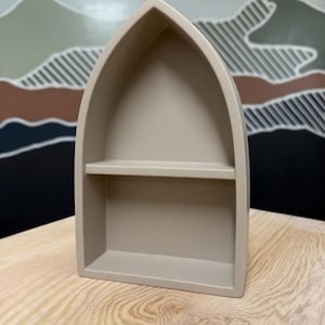 May include: A beige, pointed-arch shelf with a single horizontal divider. The structure features a smooth, matte finish and a hollow interior, creating two distinct display levels within the frame of the arch.