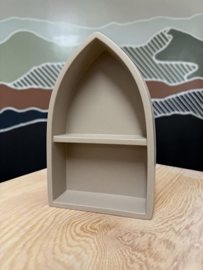 May include: A beige, pointed-arch shelf with a single horizontal divider. The structure features a smooth, matte finish and a hollow interior, creating two distinct display levels within the frame of the arch.