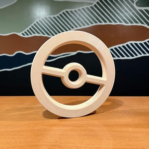 Op de afbeelding: Een lichtbeige, cirkelvormig, driedimensionaal object met een centrale ring verbonden door een horizontale balk, wat doet denken aan het ontwerp van een gestileerde bol. Het item heeft een gladde, matte afwerking en een hol centrum.