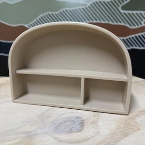 May include: A beige arch-shaped wall shelf featuring a smooth, matte finish. The structure includes a curved top frame, a single horizontal shelf spanning the width, and a vertical divider that creates two distinct storage compartments below.