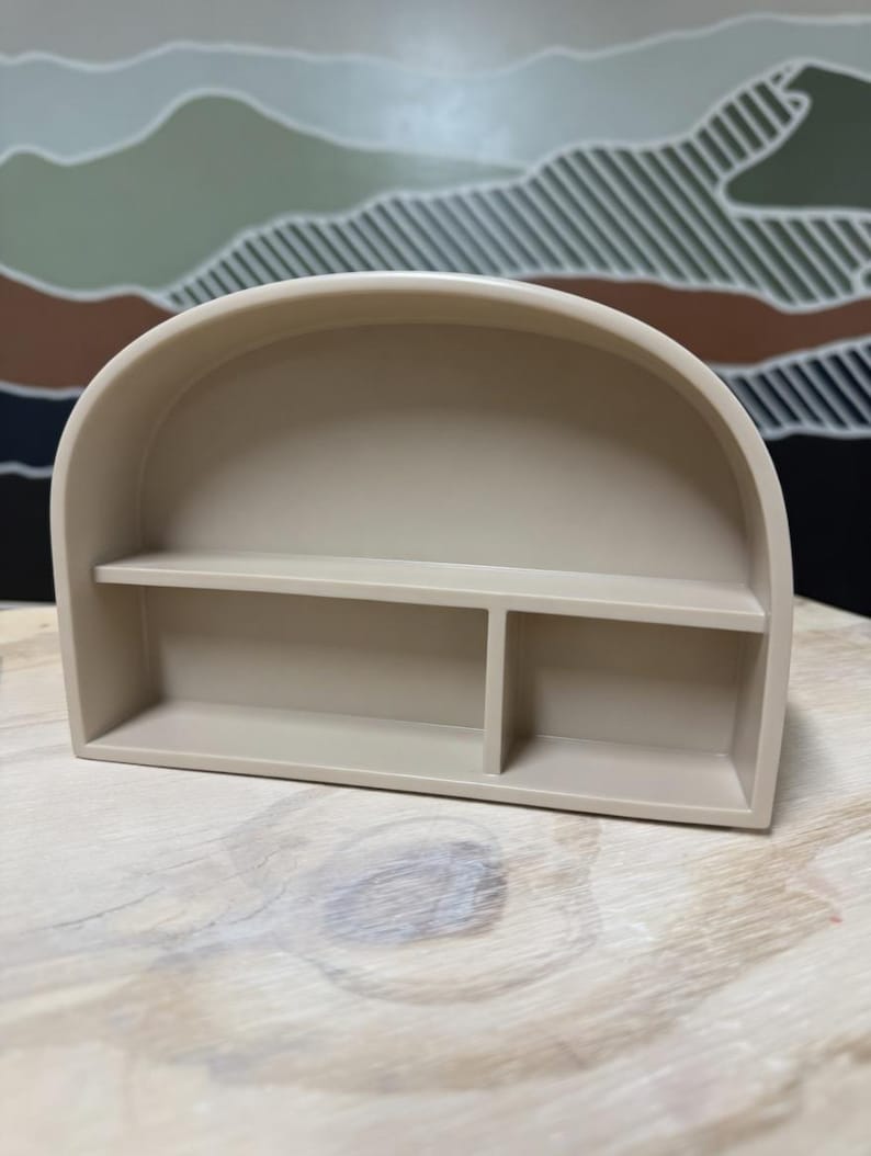 May include: A beige arch-shaped wall shelf featuring a smooth, matte finish. The structure includes a curved top frame, a single horizontal shelf spanning the width, and a vertical divider that creates two distinct storage compartments below.