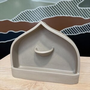 May include: A matte beige ceramic shelf with a pointed arch top and a small semi-circular ledge protruding from the center. The structure features a flat base and a smooth, uniform finish, designed as a wall-mounted or tabletop display piece.