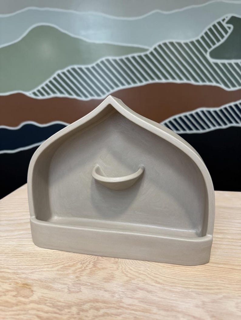 May include: A matte beige ceramic shelf with a pointed arch top and a small semi-circular ledge protruding from the center. The structure features a flat base and a smooth, uniform finish, designed as a wall-mounted or tabletop display piece.