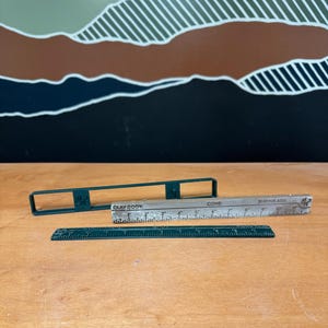 Könnte beinhalten: Ein Set aus drei Töpferwerkzeugen, bestehend aus einem dunkelgrünen rechteckigen Rahmen, einem silberfarbenen Schrumpfmaßstab mit der Aufschrift CLAY BODY, CONE und SHRINKAGE sowie einem dunkelgrünen Lineal bis 30 cm für Ton.