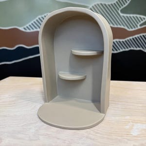 May include: A beige arched display shelf featuring a smooth, matte finish with a rounded top frame. The structure includes a flat base and two small, semi-circular shelves positioned at different heights along the interior back wall of the unit.
