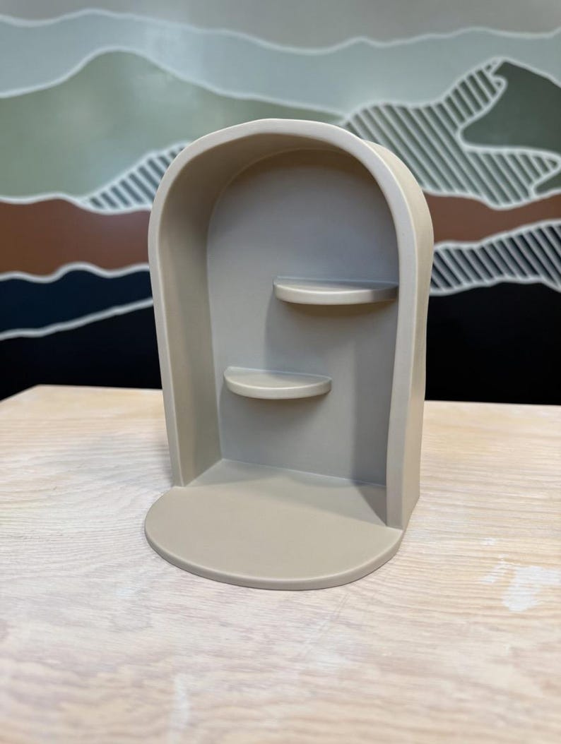 May include: A beige arched display shelf featuring a smooth, matte finish with a rounded top frame. The structure includes a flat base and two small, semi-circular shelves positioned at different heights along the interior back wall of the unit.