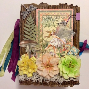 May include: A handmade journal with a fairy theme. The cover is made of corrugated cardboard and decorated with paper flowers, a dragonfly, and a fairy illustration. Colorful ribbons and decorative stamps add to the whimsical design.