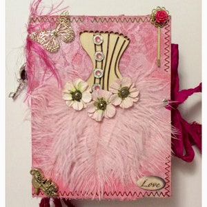 May include: Pink journal with a pink feather design, a wooden corset cutout, and a pink flower embellishment. The journal has a pink ribbon and the word "Love" written on the front.