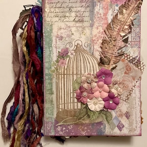 May include: A purple and white journal with a birdcage and flowers on the cover. The journal has a decorative tassel made of colorful ribbons.